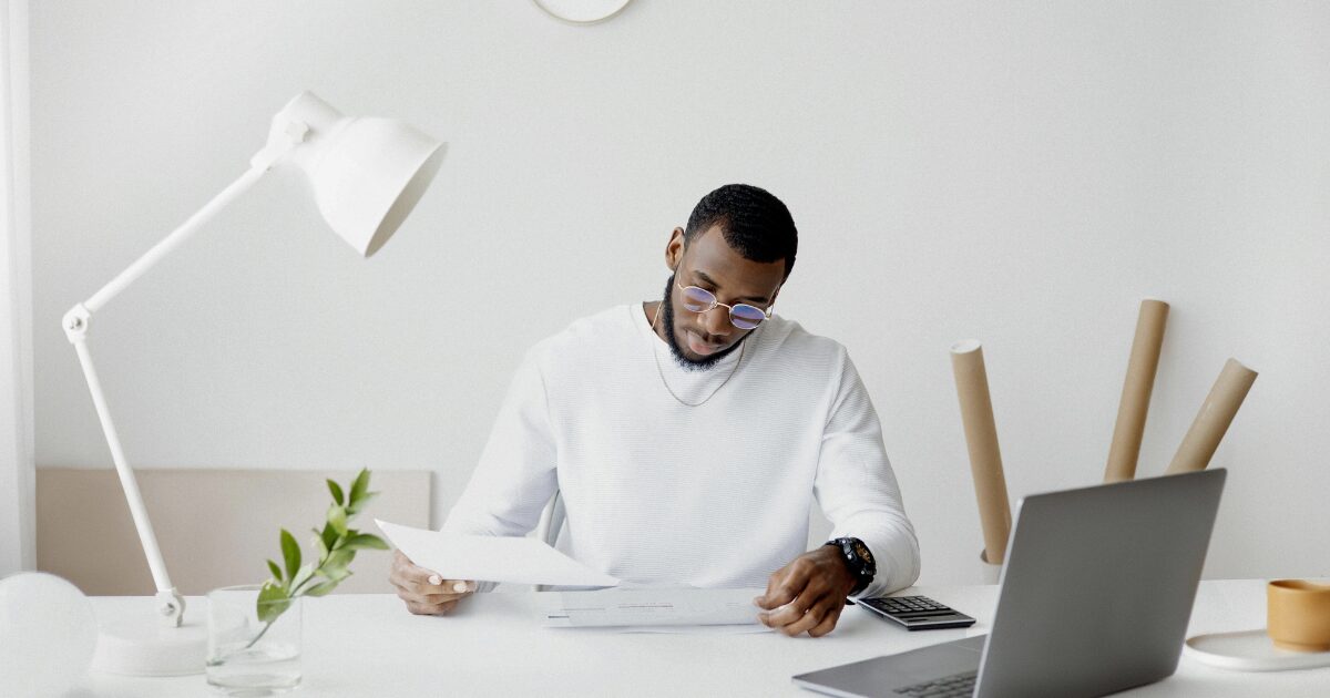 A Black man wearing a white long-sleeve shirt, seated in front of an open laptop while reading documents.