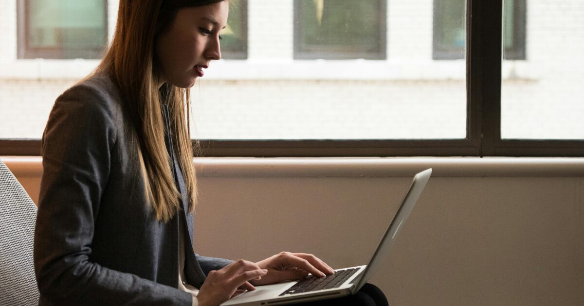 A Caucasian woman wearing a black blazer uses a laptop on her lap.