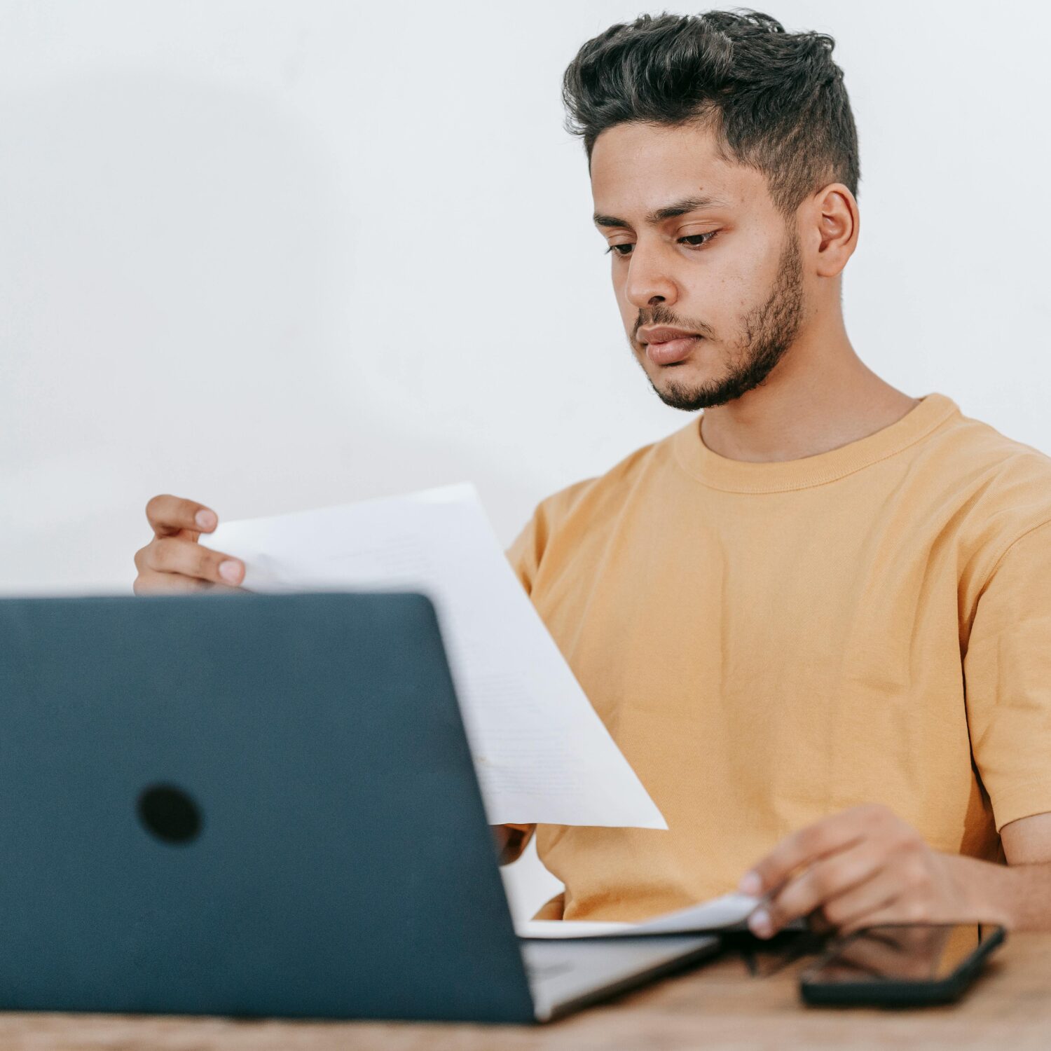 A South Asian man wearing a light orange t-shirt reads a document while sitting in front of an open laptop