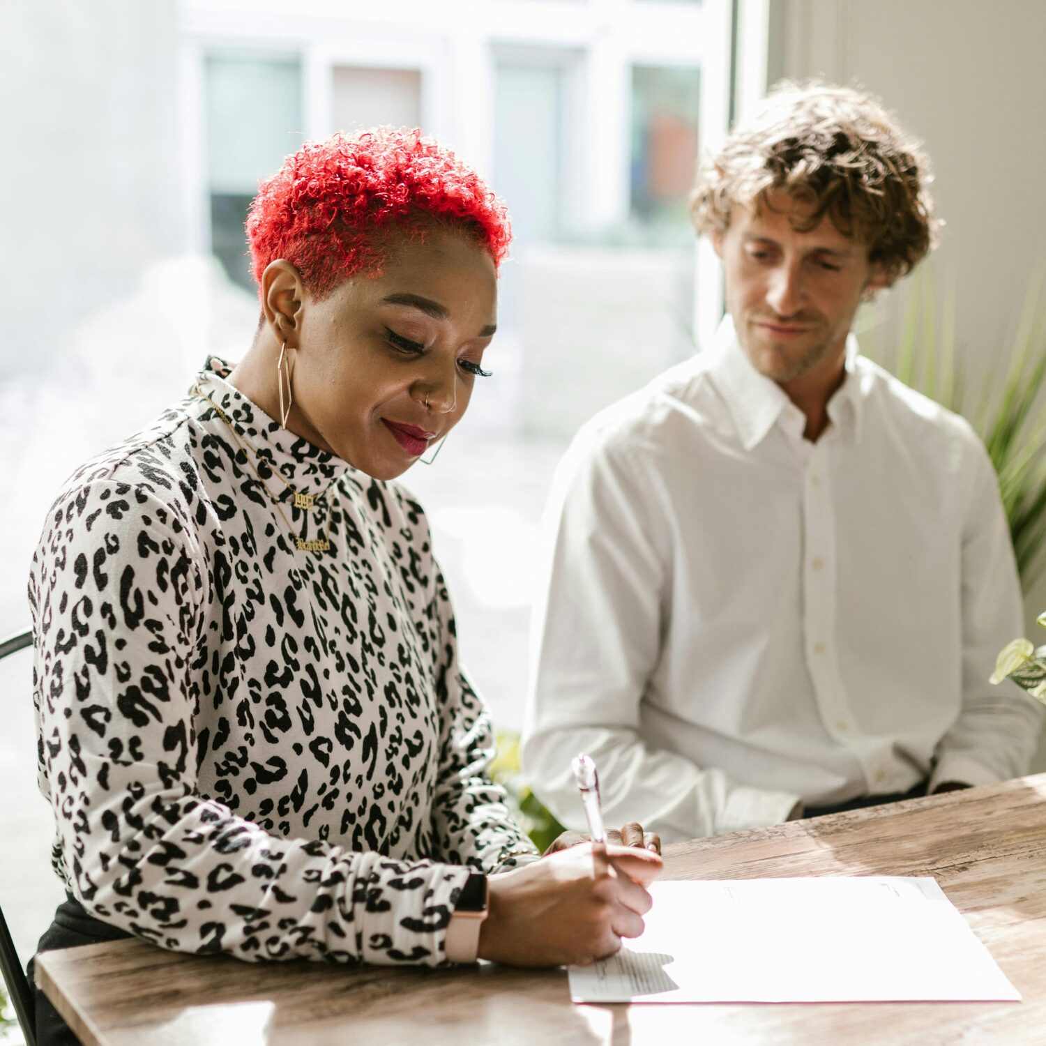 A Black woman wearing an animal-print blouse writes on a document while a Caucasian man wearing a white long-sleeve shirt watches her write.