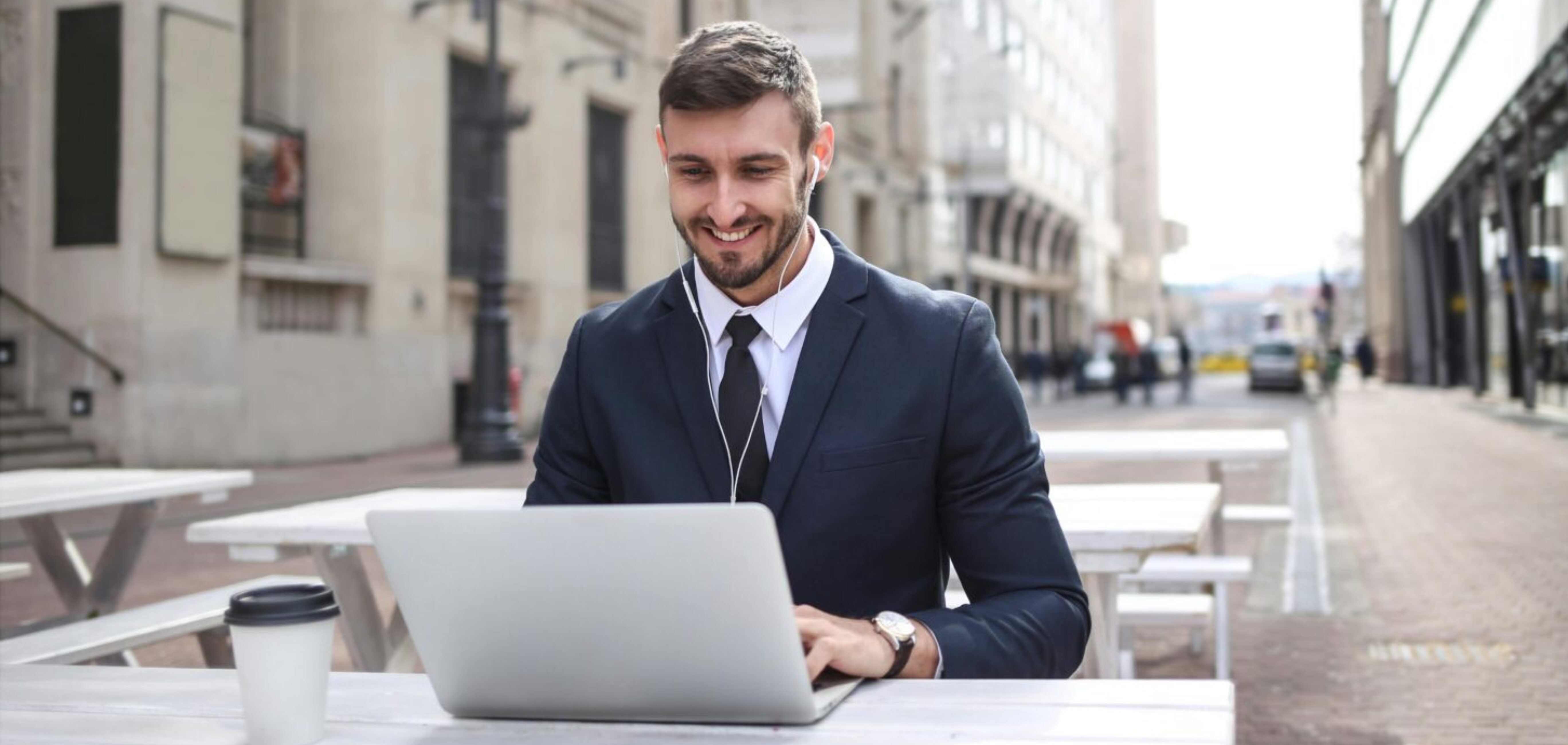 A Caucasian man in a navy suit sitting on a patio on a city street, with earphones in his ears and an open laptop on the table.