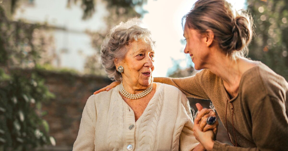 417 An elderly Caucasian woman wearing a cream-coloured cardigan smiles while holding hands with a Caucasian woman wearing a brown shirt, who rests her right hand on the elderly woman’s back.