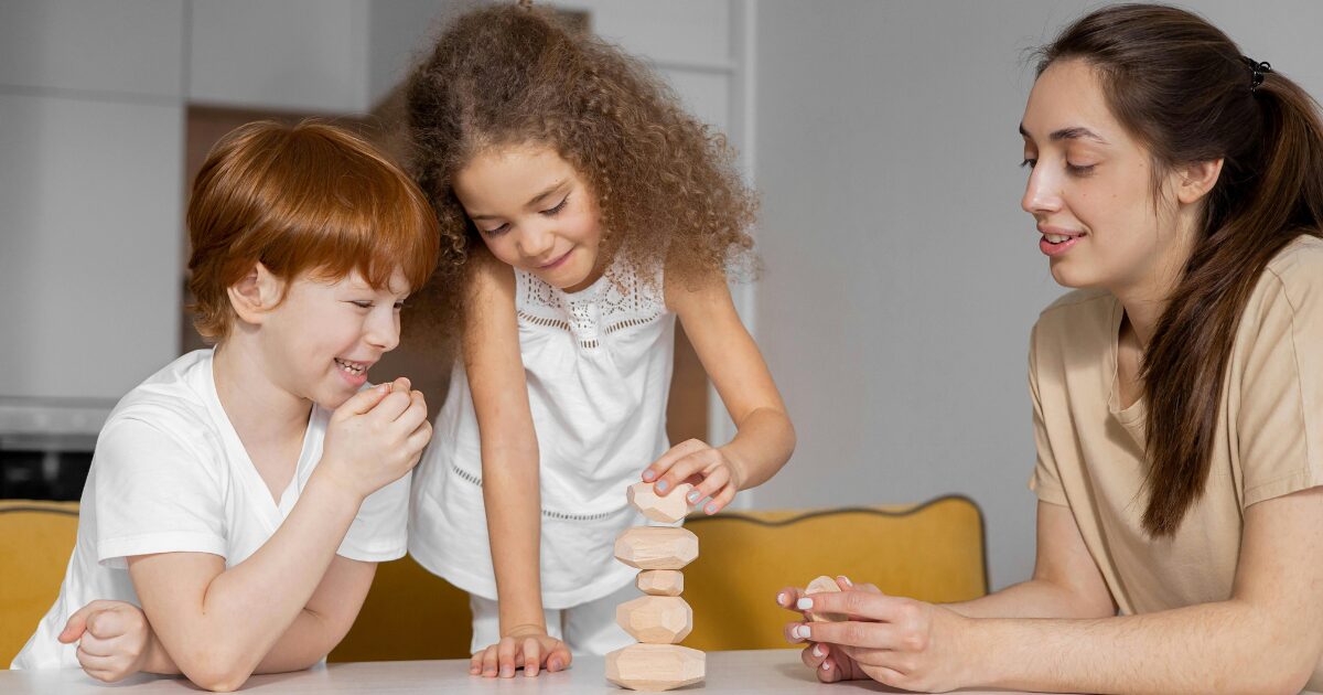 418 A Caucasian woman wearing a tan shirt watches a young Caucasian boy and girl playing with building blocks on a table.