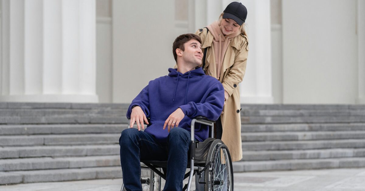 420 A Caucasian man wearing a blue hooded jacket sits in a wheelchair while a Caucasian woman wearing a light brown coat pushes it.