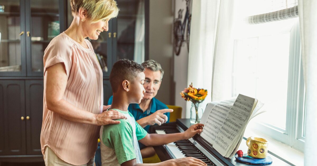 421 A young Black boy sits in front of a piano while a Caucasian woman stands behind him, resting her hands on his back, and a Caucasian man beside him points to a music sheet.