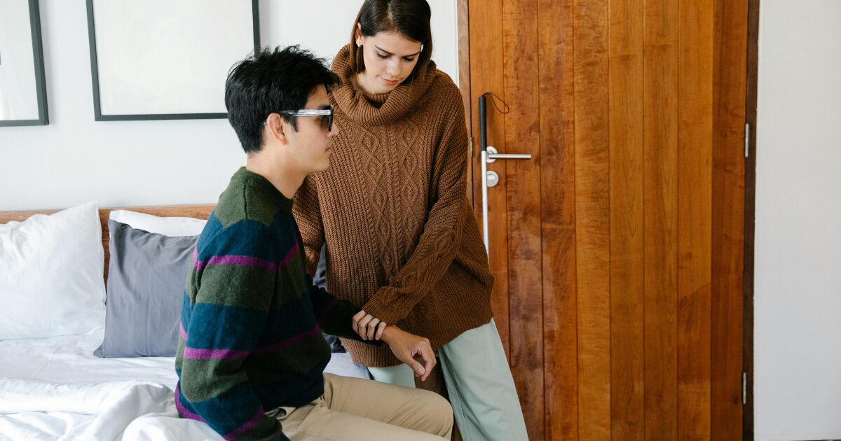422 A Caucasian man wearing a striped sweater sits on a bed while a Caucasian woman wearing a brown sweater stands beside him, assisting by holding his arm.