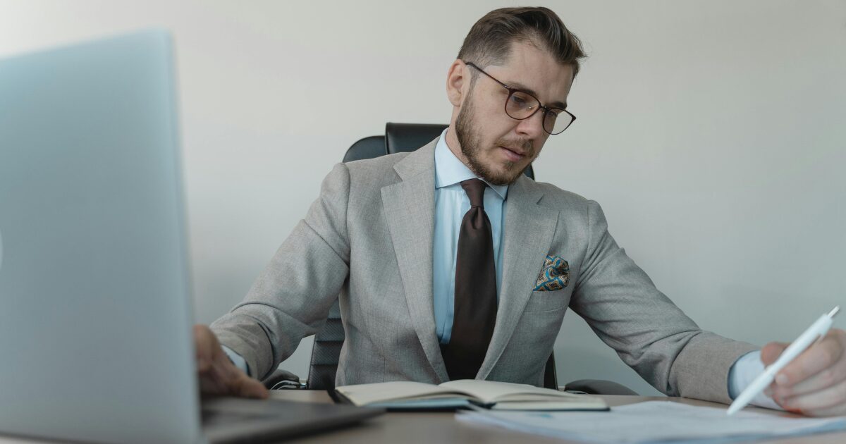 423 A Caucasian man wearing a light grey business suit writes on a document with his left hand while typing on a laptop with his right hand.