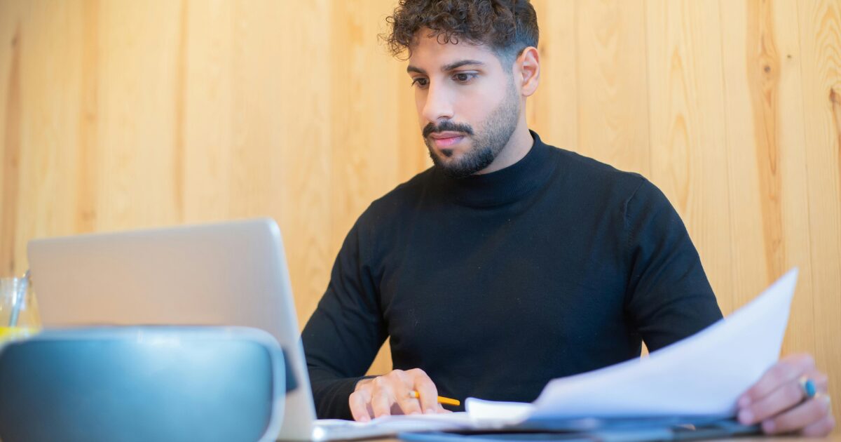 425 A Caucasian man wearing a black long-sleeve shirt looks at a laptop screen while holding documents in his left hand.