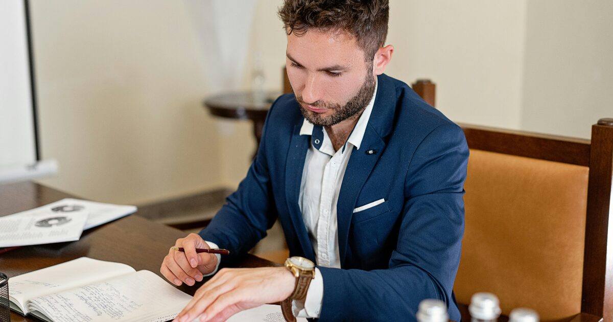 426 A Caucasian man wearing a dark blue business suit reads from a notebook while holding a pen in his right hand.