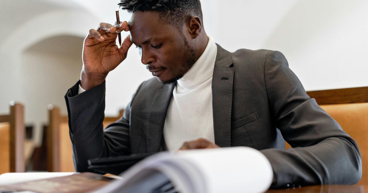 427 A Black man wearing a dark grey business suit touches his forehead with his right hand holding a pen as he reads a document.