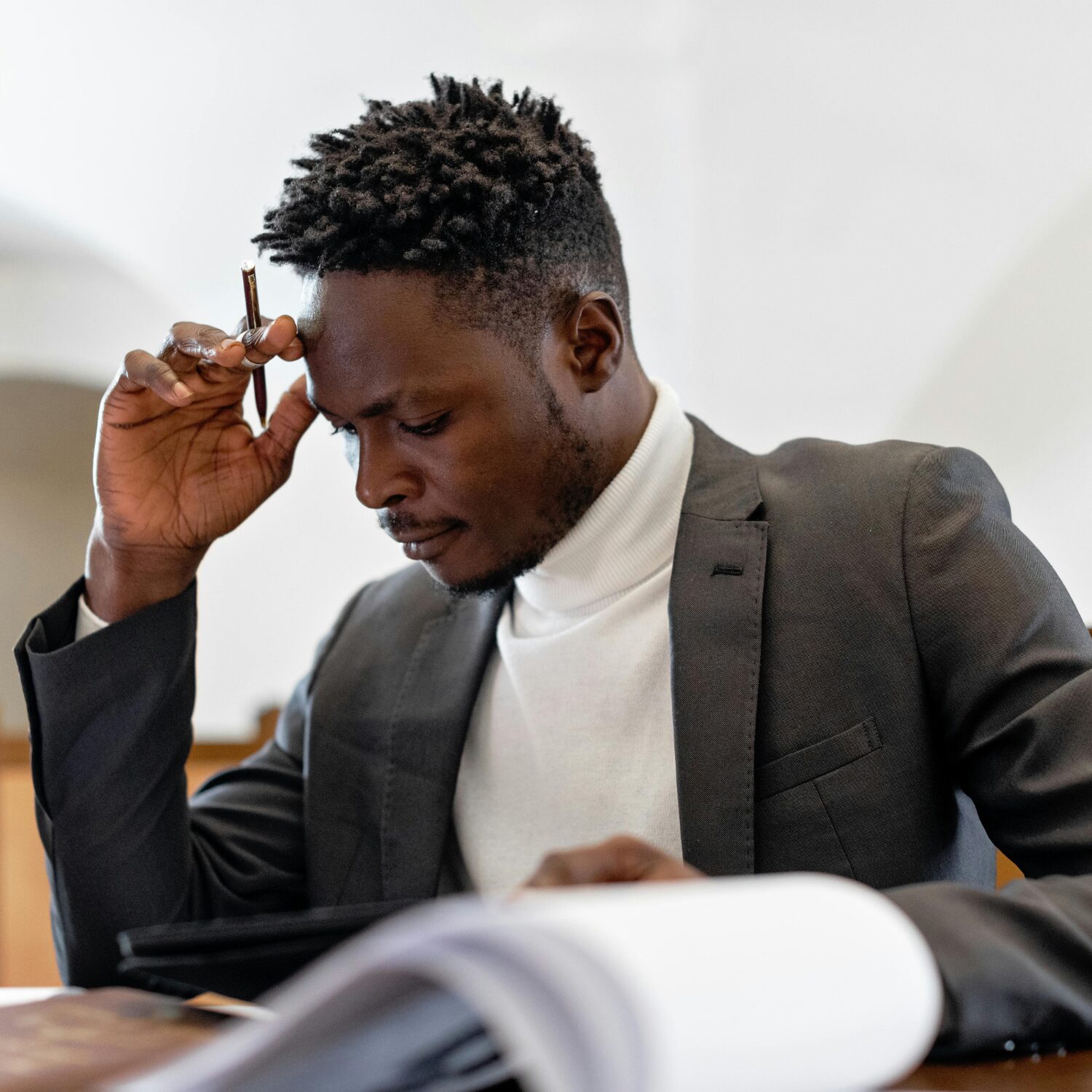 A Black man wearing a dark grey business suit touches his forehead with his right hand holding a pen as he reads a document.