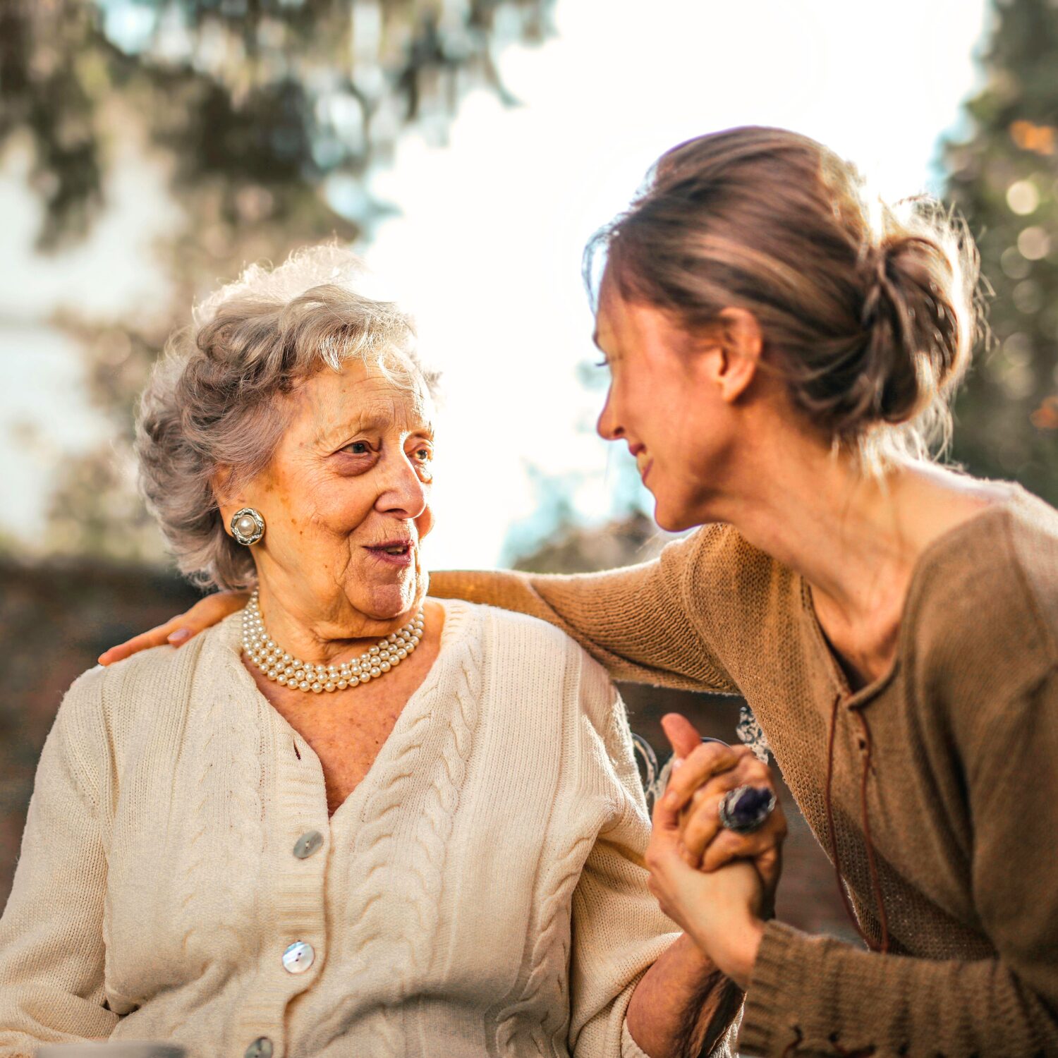 An elderly Caucasian woman wearing a cream-coloured cardigan smiles while holding hands with a Caucasian woman wearing a brown shirt, who rests her right hand on the elderly woman’s back.