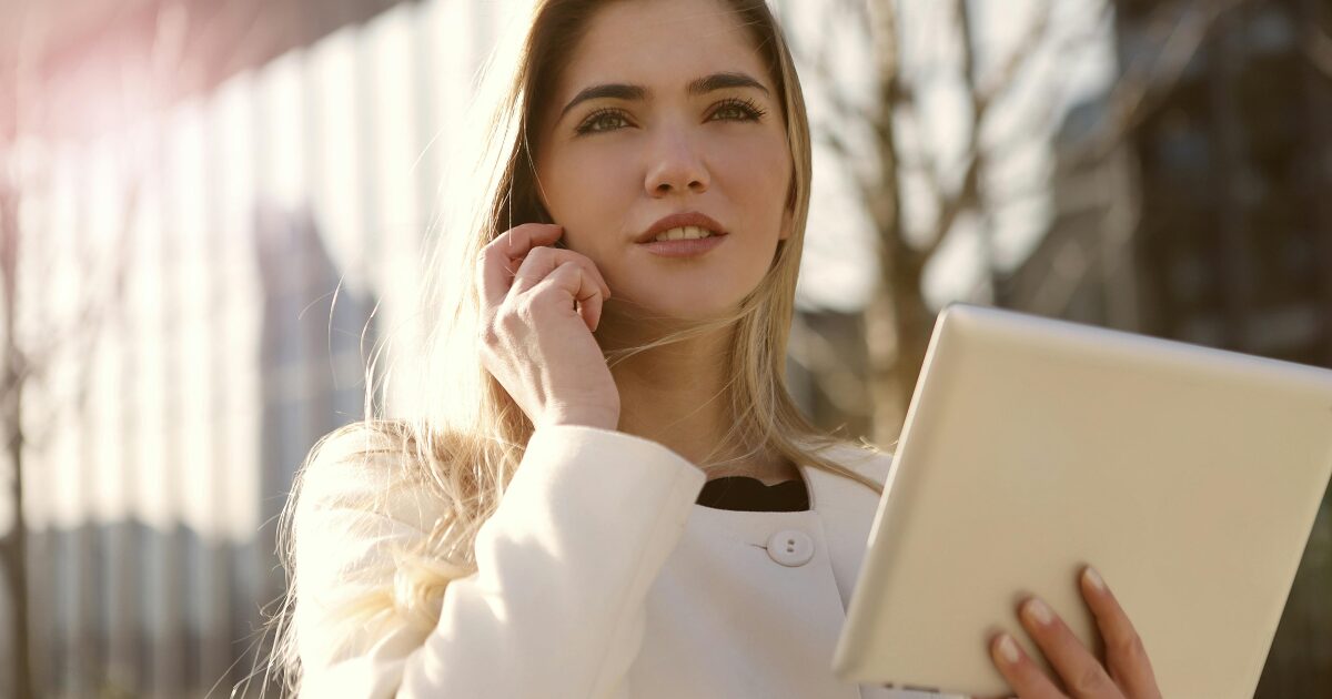 429 A Caucasian woman wearing a white long-sleeve shirt holds a phone to her ear while holding a tablet in her left hand.