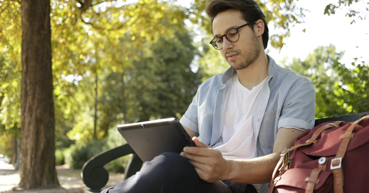 431 A Caucasian man wearing a light blue denim shirt sitting outdoors while using a tablet.