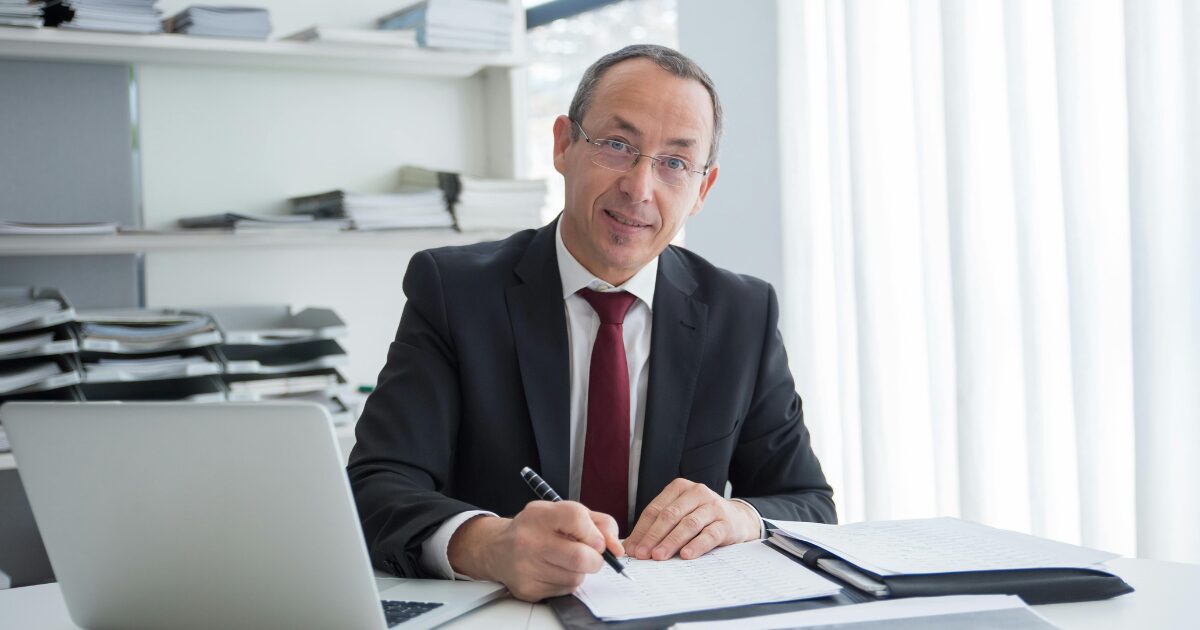 A Caucasian man in a black business suit writes on a document in a folder.