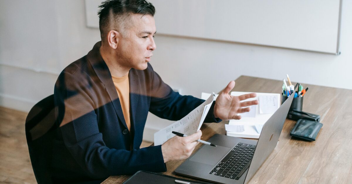 A Caucasian man wearing a dark blue suit holds a pen and documents while talking in front of a laptop.