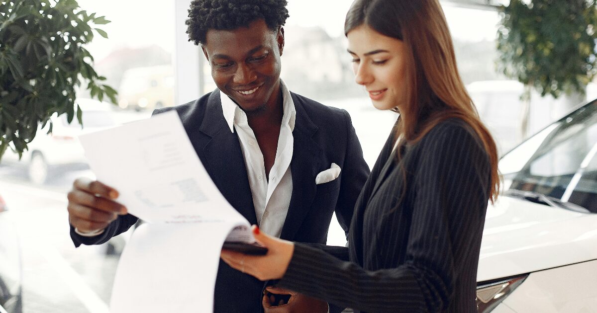 A Caucasian woman wearing a black blazer shows documents to a Black man wearing a black business suit.
