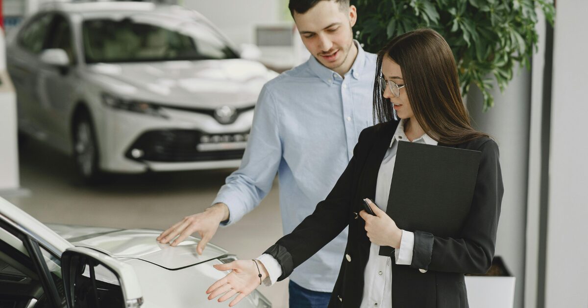 A Caucasian woman wearing a black business suit shows a white car to a Caucasian man wearing a light blue shirt.