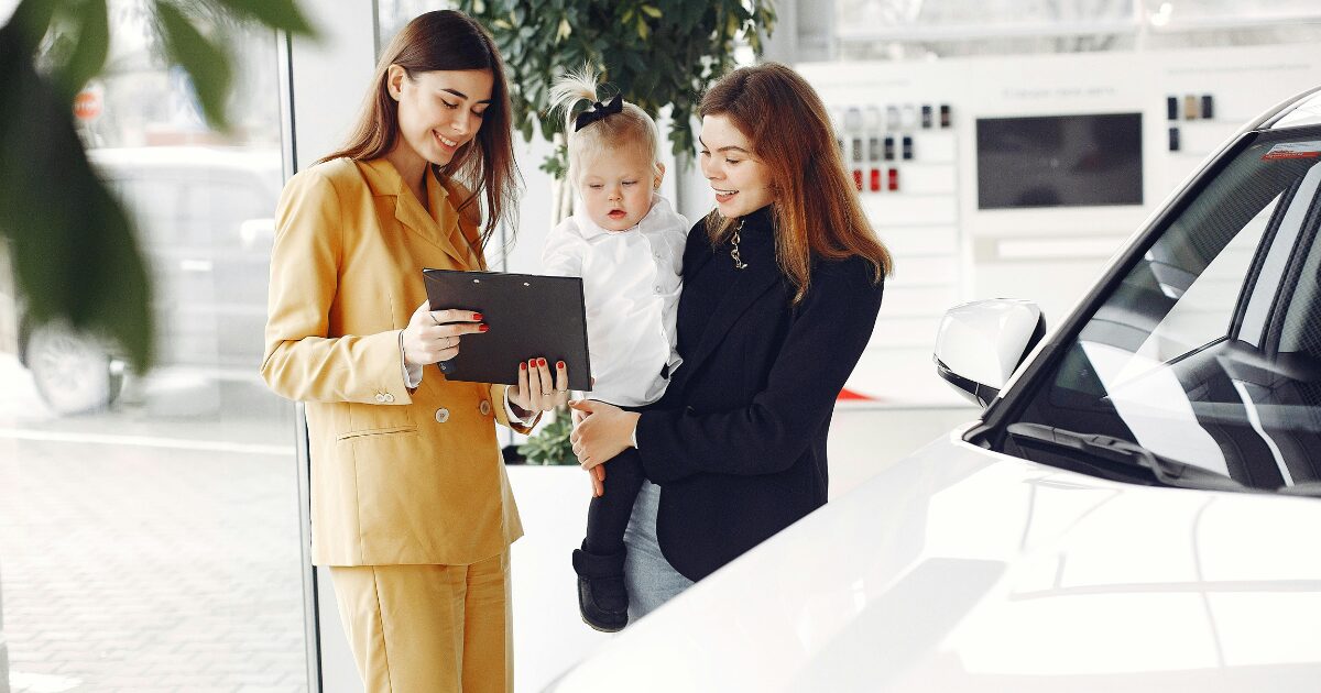 A Caucasian woman wearing a mustard coat shows a document to a Caucasian woman wearing a black coat who is carrying a Caucasian baby girl in her arms.