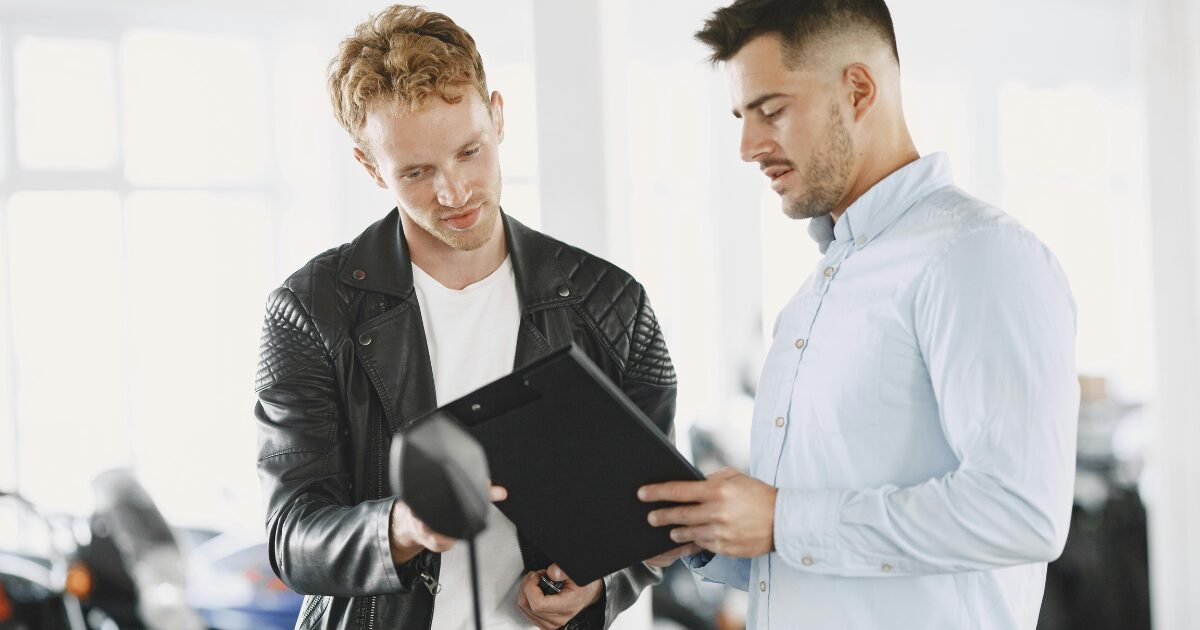 A Caucasian man wearing a white long-sleeve shirt shows documents to a Caucasian man wearing a black leather jacket.