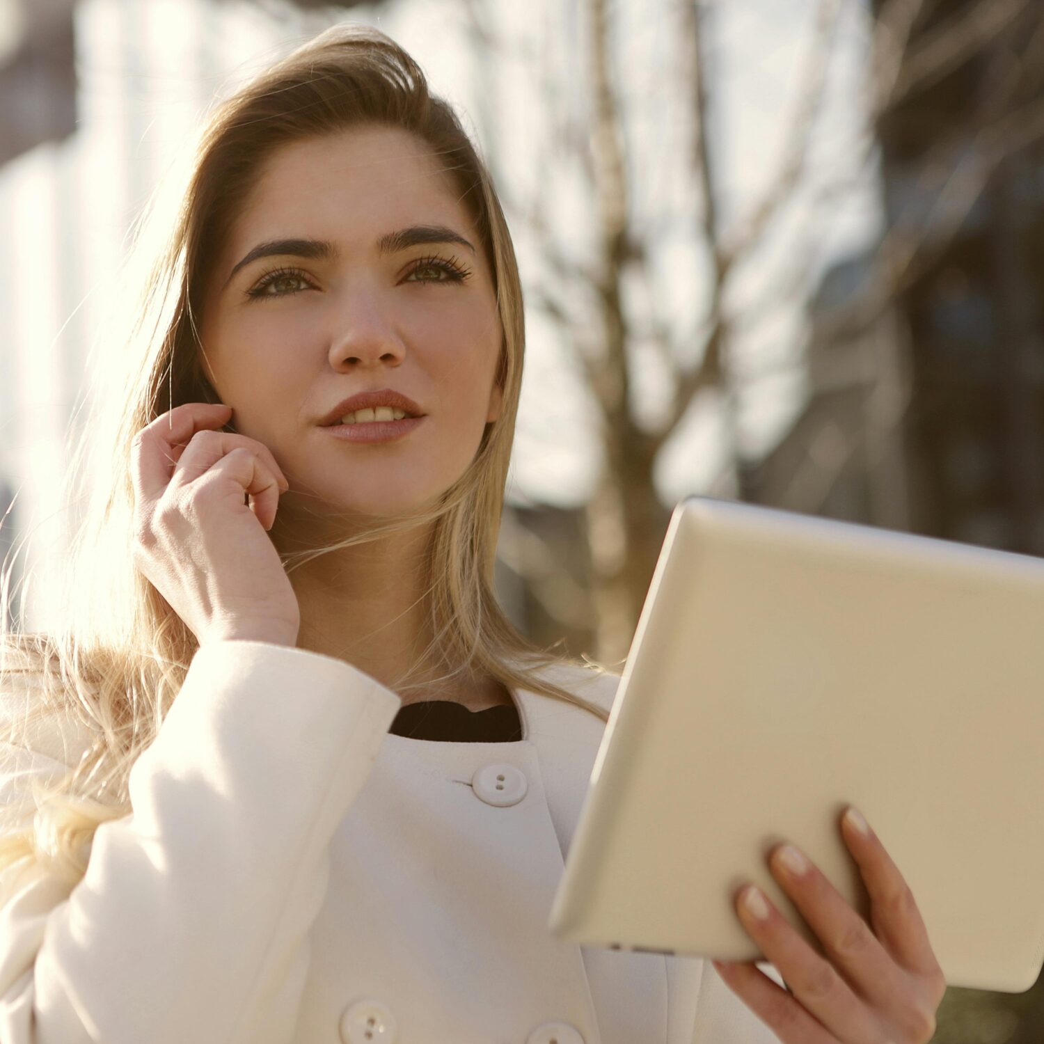 A Caucasian woman wearing a white long-sleeve shirt holds a phone to her ear while holding a tablet in her left hand.