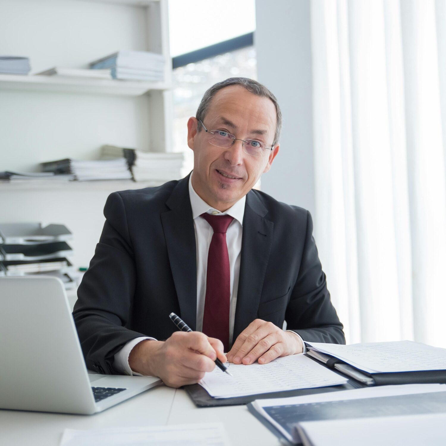 A Caucasian man in a black business suit writes on a document in a folder.
