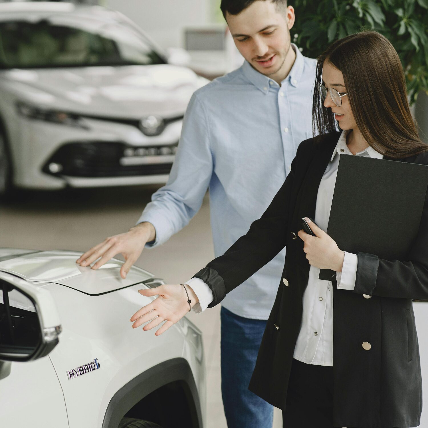 A Caucasian woman wearing a black business suit shows a white car to a Caucasian man wearing a light blue shirt.