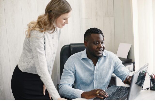 A Black man sitting at a desk with his laptop open, and a white woman is standing beside him as he shows her something on his screen. They are both dressed business casual.