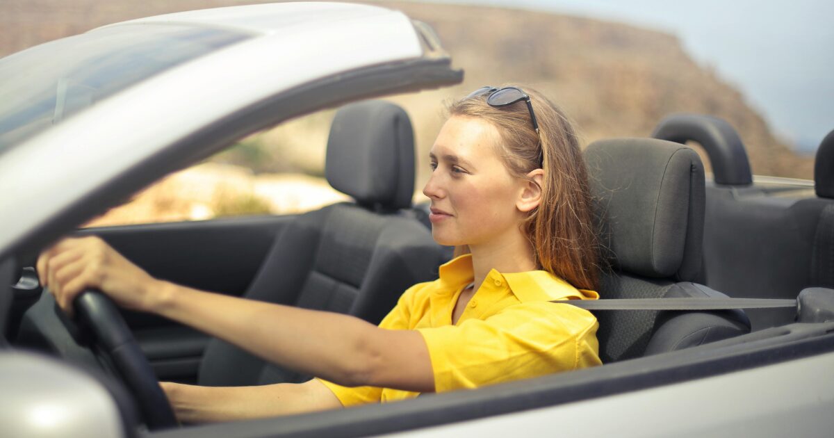 A Caucasian woman wearing a yellow shirt driving a car.
