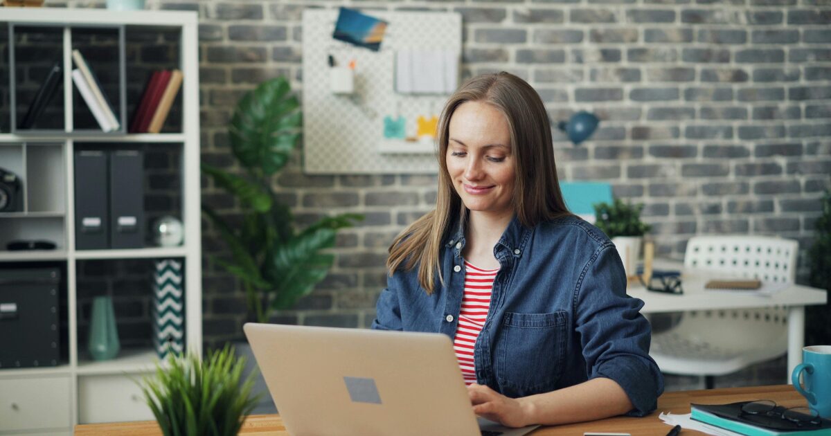 A White woman wearing a blue button-up shirt smiles while typing on her laptop.