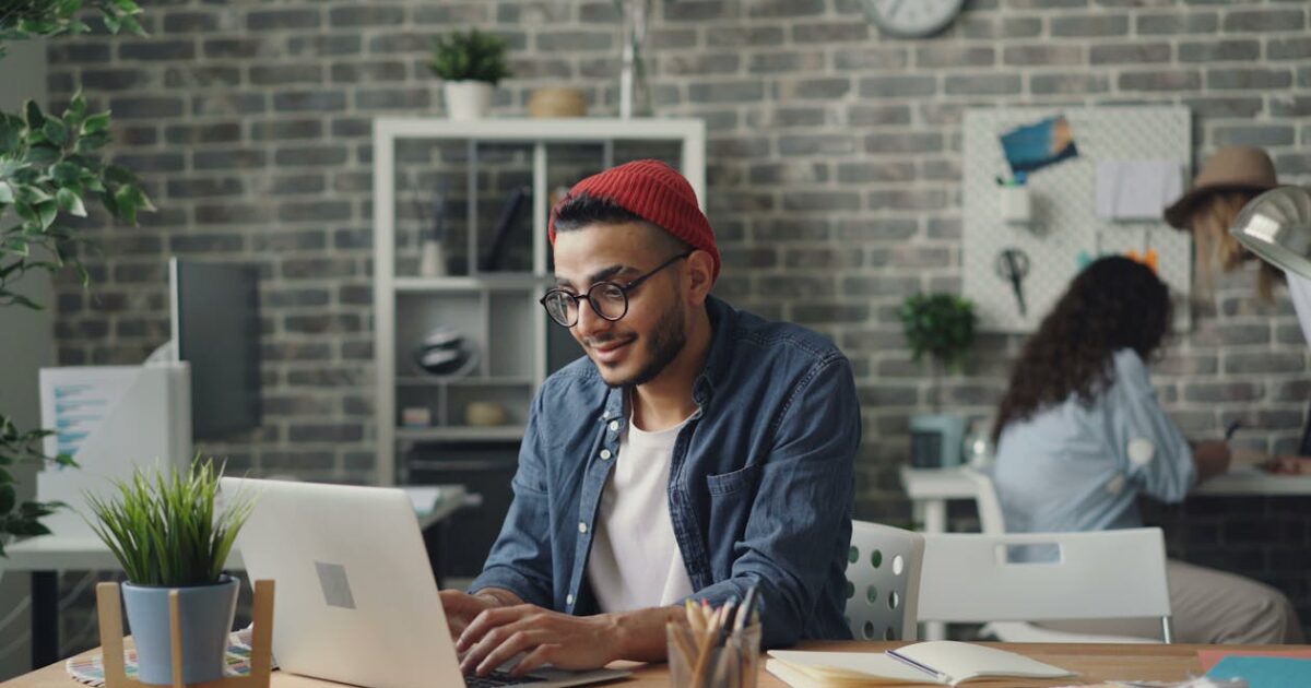 A South Asian man wearing a blue button-up shirt and eyeglasses smiles while typing on his laptop.