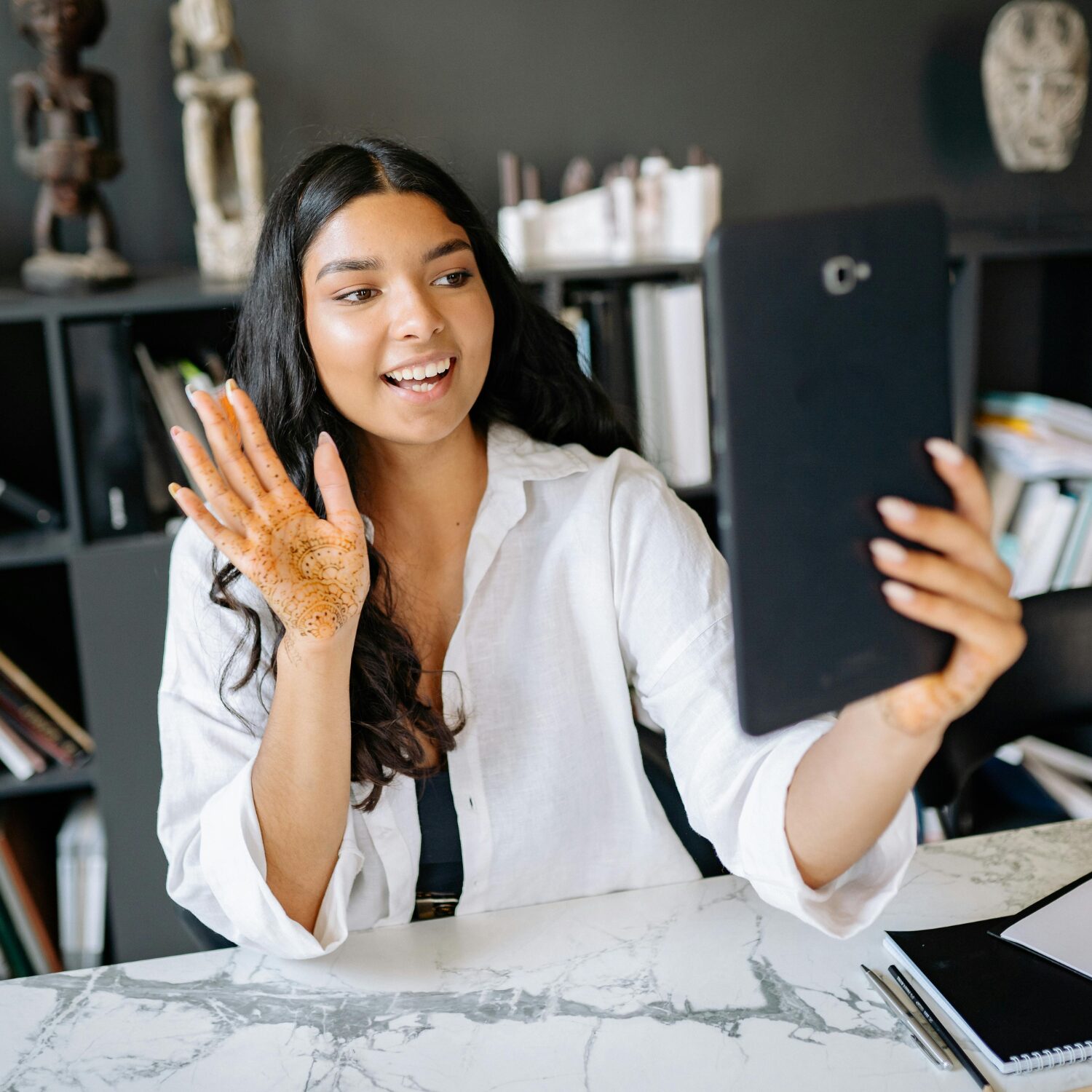 A young South Asian woman in a white blouse waving to someone on a video call on her tablet, sitting in an office.