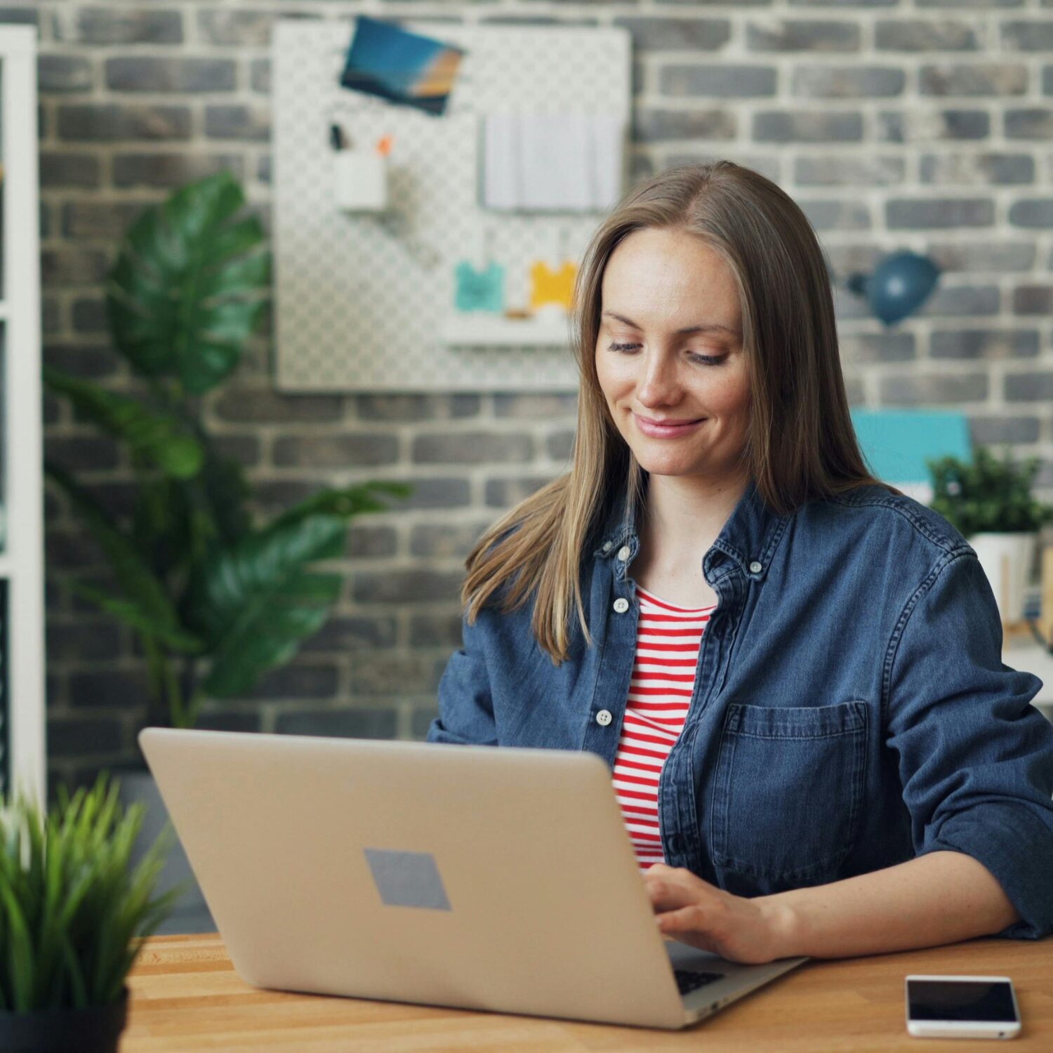 A White woman wearing a blue button-up shirt smiles while typing on her laptop.