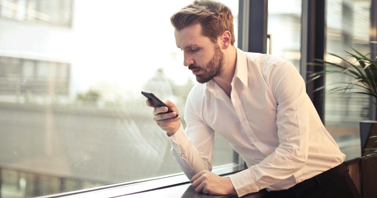 In-Blog Thumbnails White man wearing a white button-up shirt leaning on a windowsill, looking at his smartphone