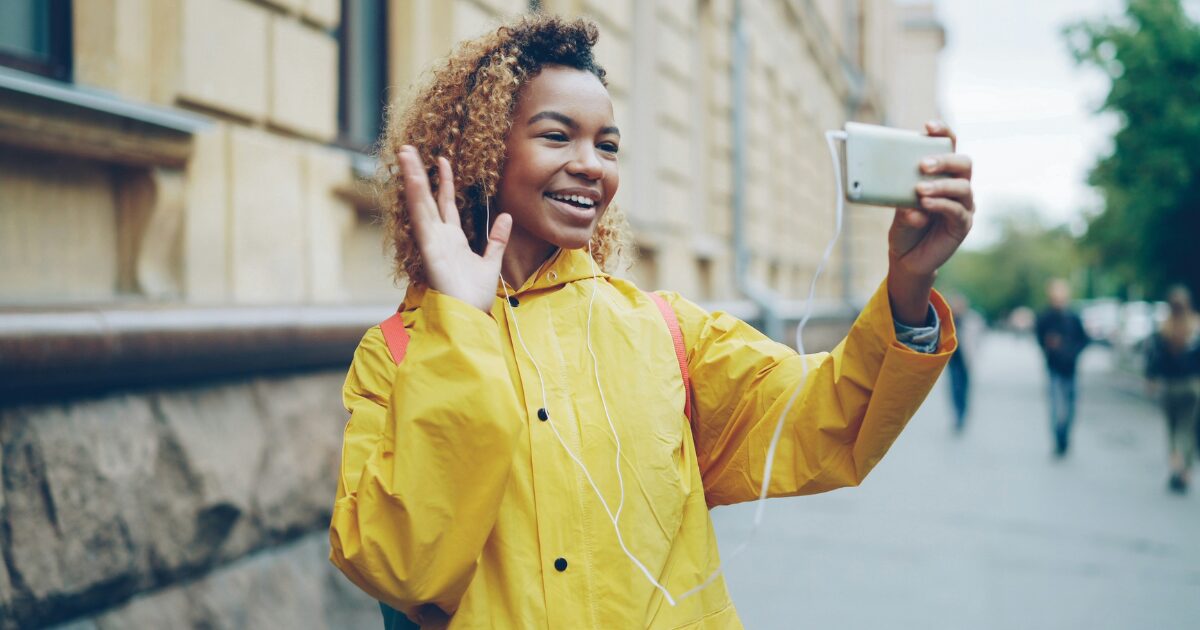 Black woman standing on the street while taking a video call from her smartphone. She's wearing a yellow raincoat and is waving to the camera.
