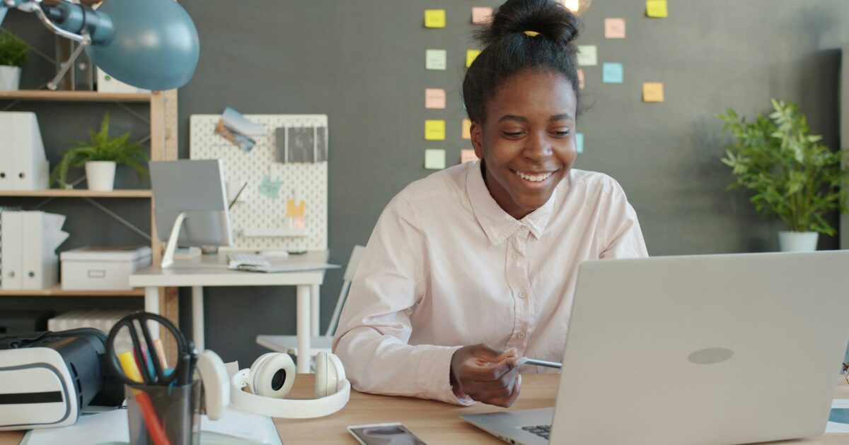 Black woman sitting in an office, she's smiling as she looks at the laptop in front on her. She's wearing a white button-up shirt.