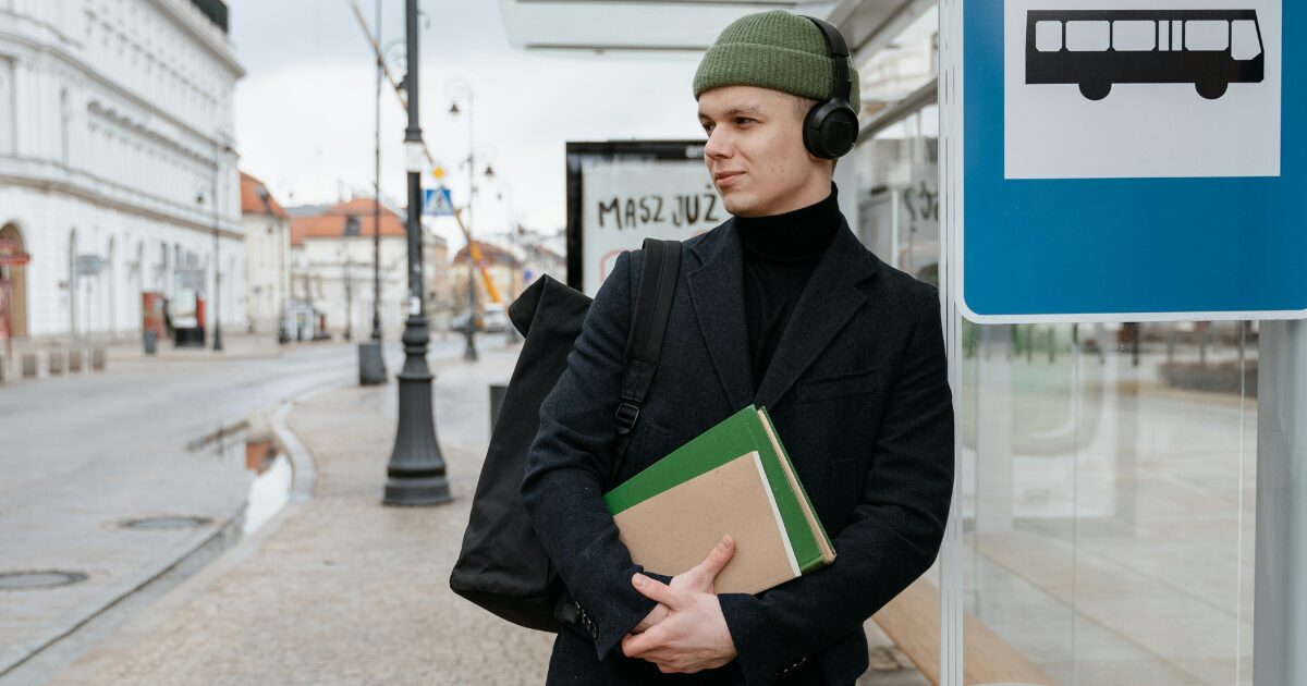 Young Caucasian man standing at a bus stop holding documents, wearing a black blazer.
