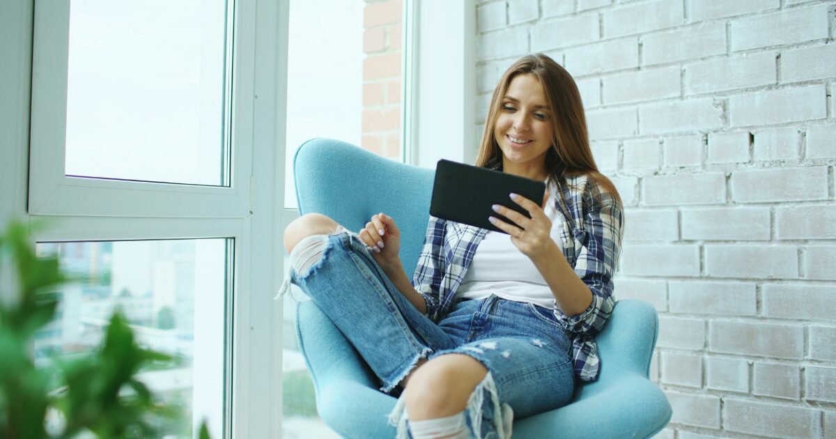 Caucasian woman sitting in a chair by a window, while smiling and holding a tablet. She's wearing ripped jeans and a plaid shirt.