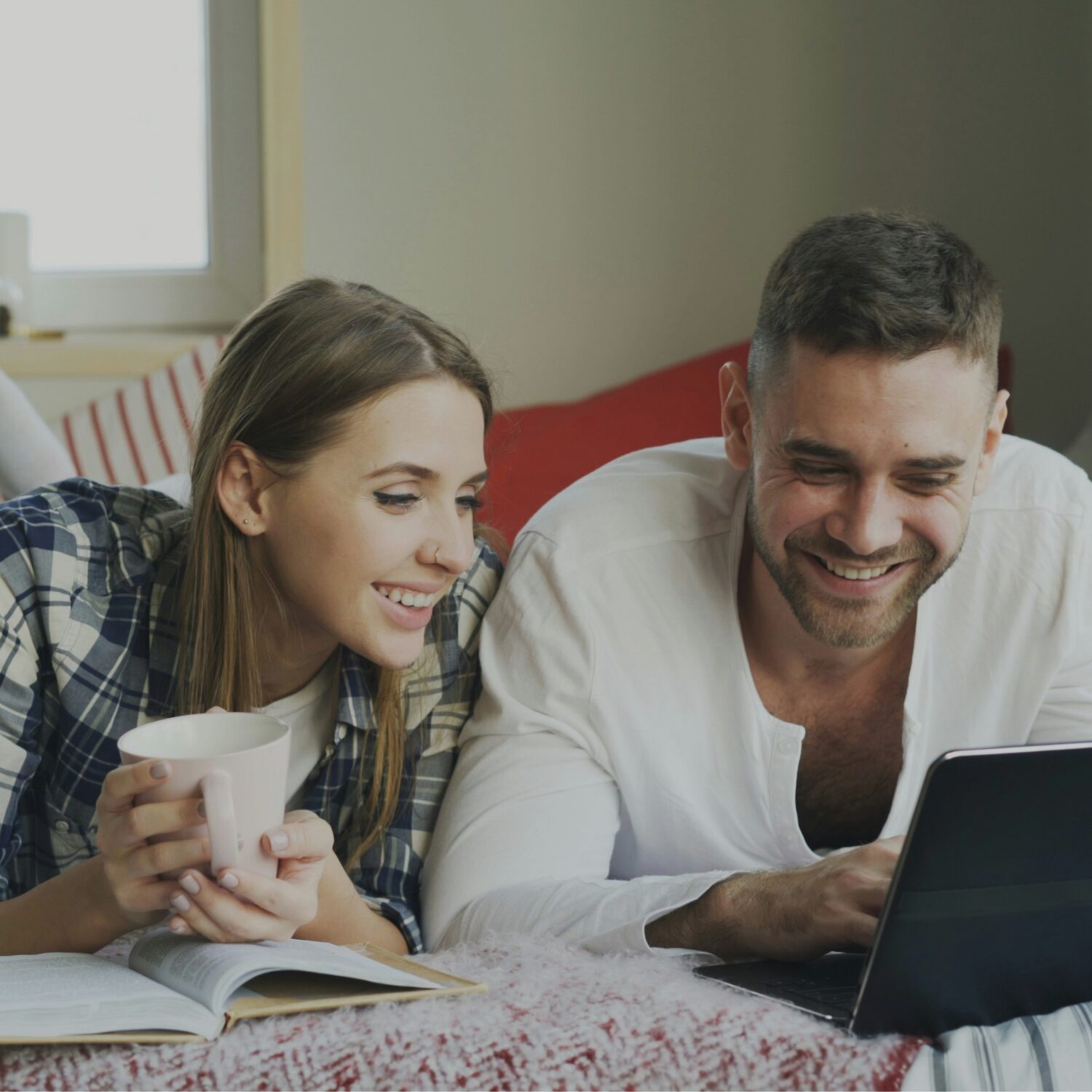 A Caucasian female and male laying on a bed and looking at a laptop in front of a male while smiling.