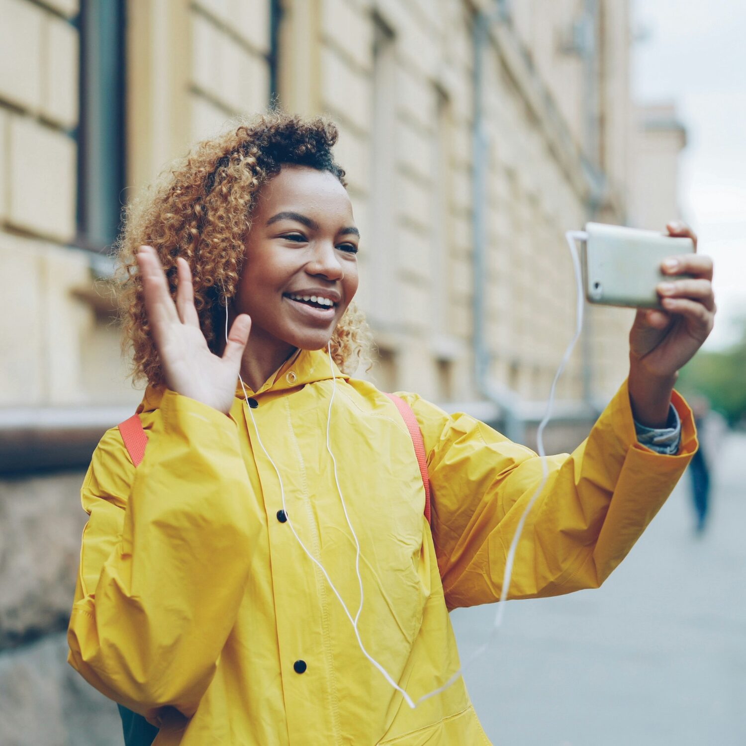 Black woman standing on the street while taking a video call from her smartphone. She's wearing a yellow raincoat and is waving to the camera.