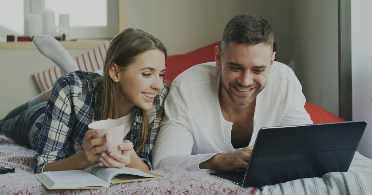 A Caucasian female and male laying on a bed and looking at a laptop in front of a male while smiling.
