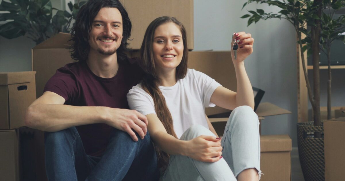 A Caucasian young male and female sitting on the floor with boxes all around them, as they smile, she's holding a key.