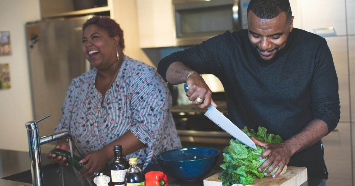 A Black woman and man in the kitchen, laughing together while cooking.