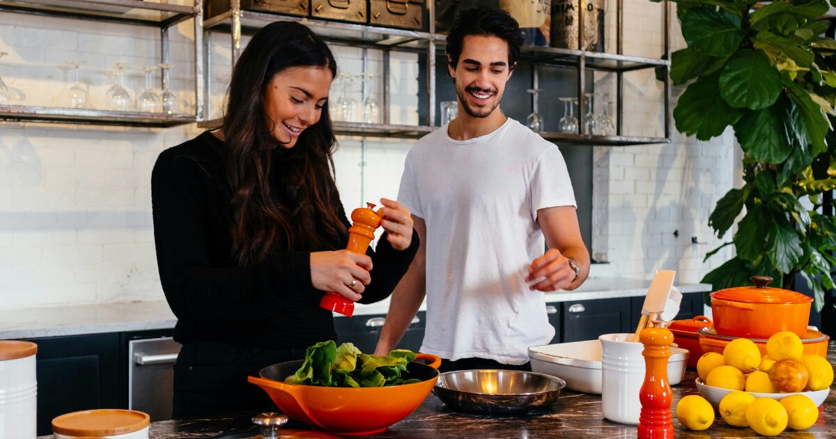 A young Latin American man and woman smiling as they cook together in a kitchen.
