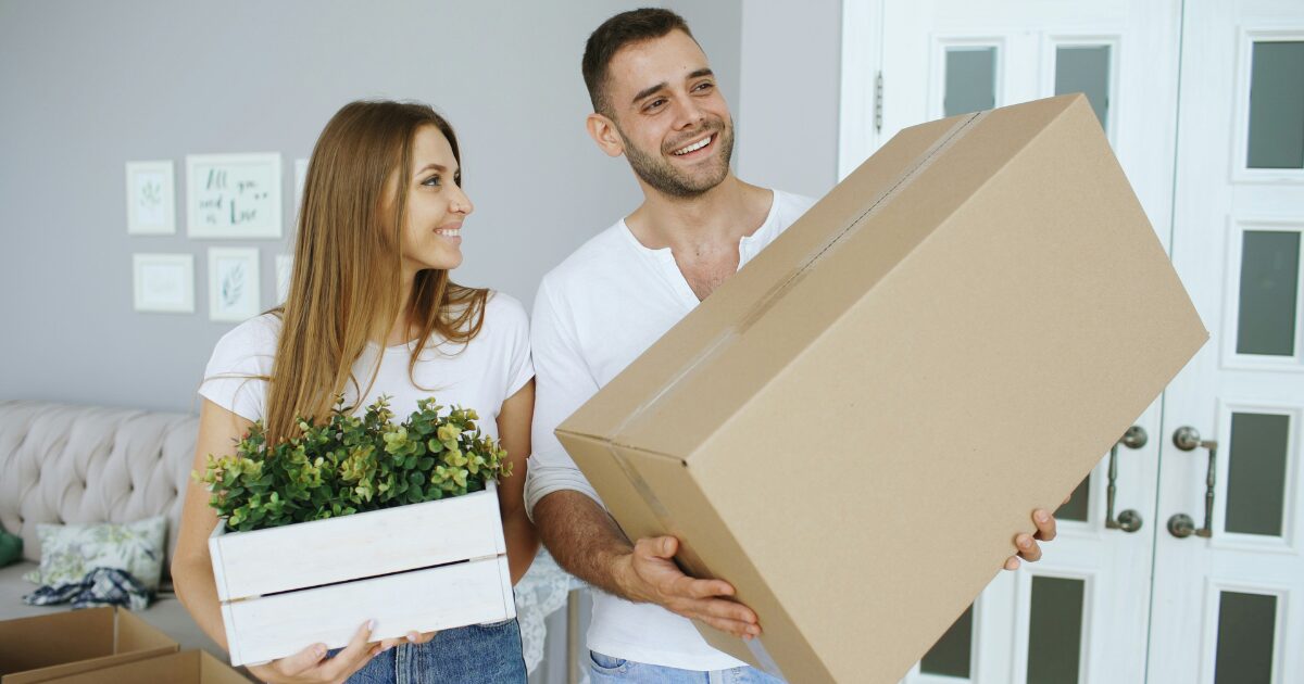 A young Caucasian male and female in a home, he is holding a moving box and she is holding a plant.