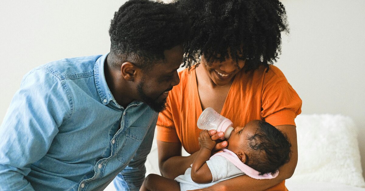 A Black man and woman smiling as they look down at a baby, who the woman is holding.