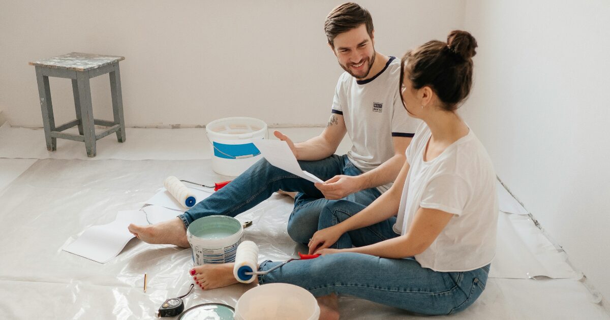 A Caucasian male and female wearing jeans and white t-shirts as they sit on the floor surrounded by painting supplies.