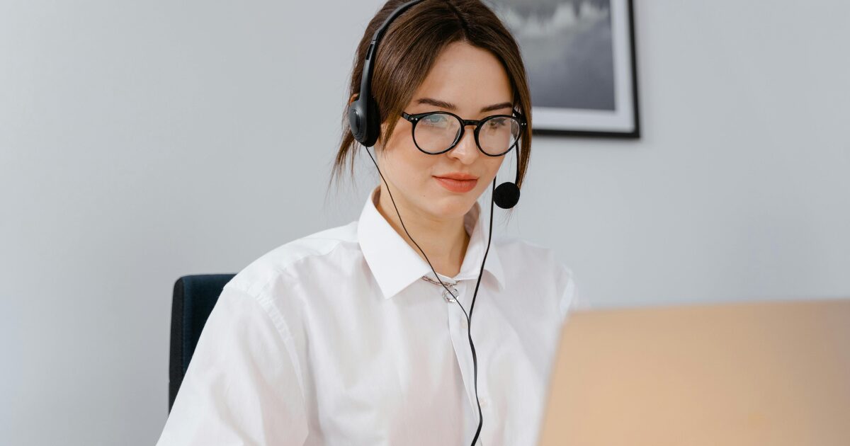 A young Caucasian woman sitting in front of an open laptop with a headset on.