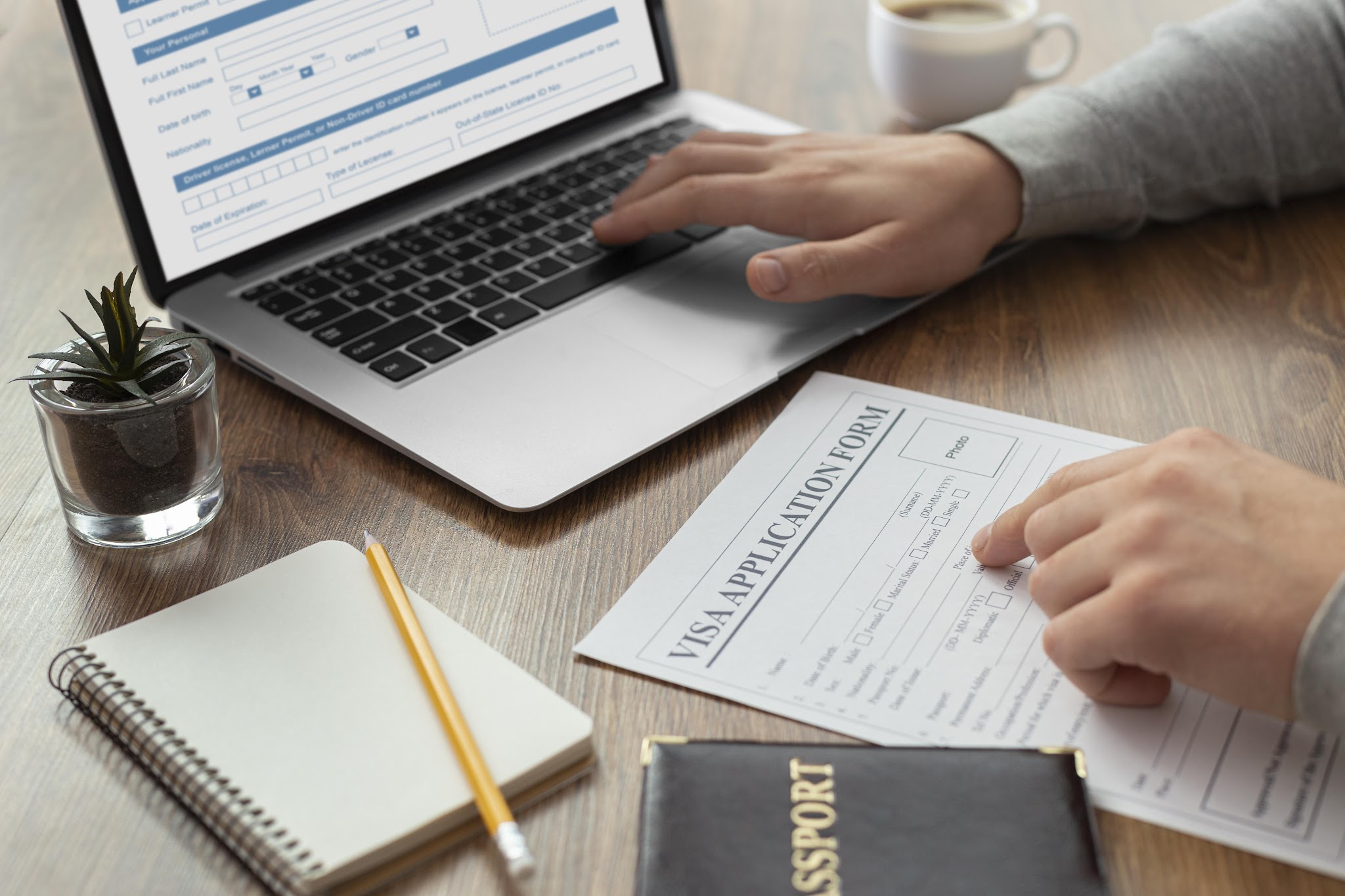 A man preparing a visa application on a laptop with a pad of paper a pencil and plant sitting on his desk