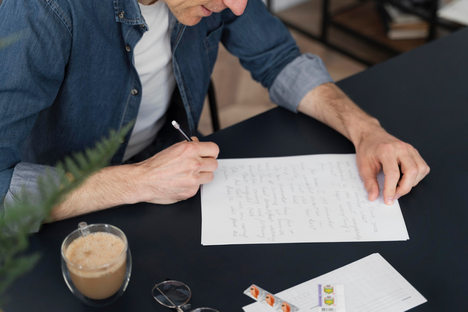 a man writing a letter on a black desk with a pencil and a cup of coffee